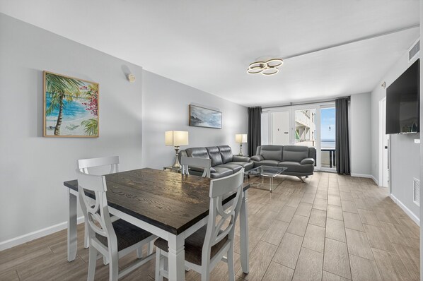 A modern living room with a dining table, white chairs, a gray sofa, and a large window with a view.
