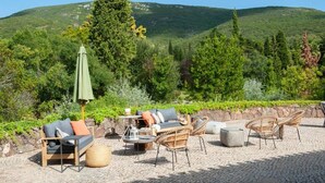 Een terras met rieten meubilair, een ronde tafel en een groene parasol, met op de achtergrond weelderige begroeiing en heuvels.