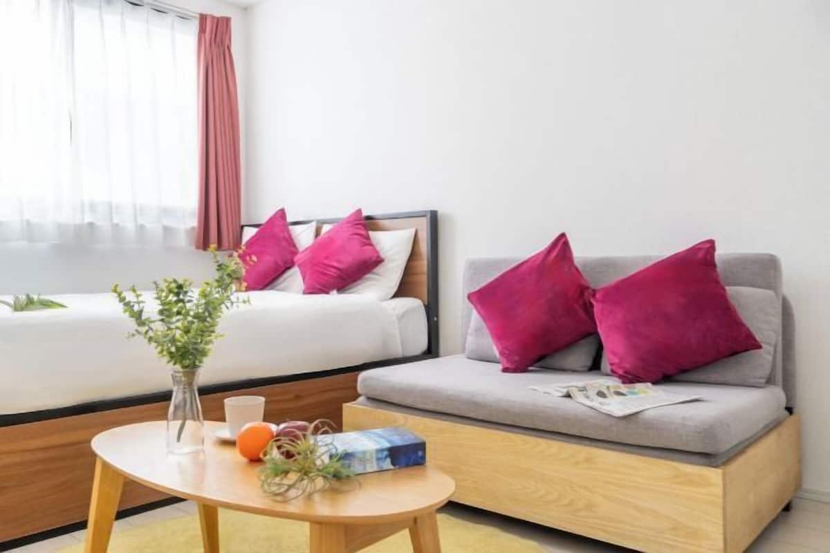 Moderni hotellihuone, jossa on harmaa sohva, puinen sohvapöytä ja vaaleanpunaisilla tyynyillä varustettu sänky.