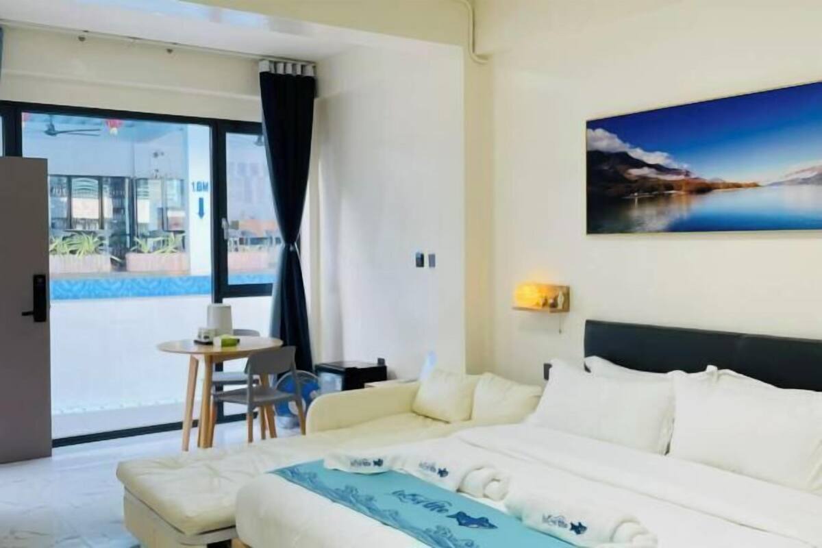 Ein Hotelzimmer mit Bett, einem kleinen Tisch mit Stuhl, einem Bild an der Wand und Blick ins Freie.