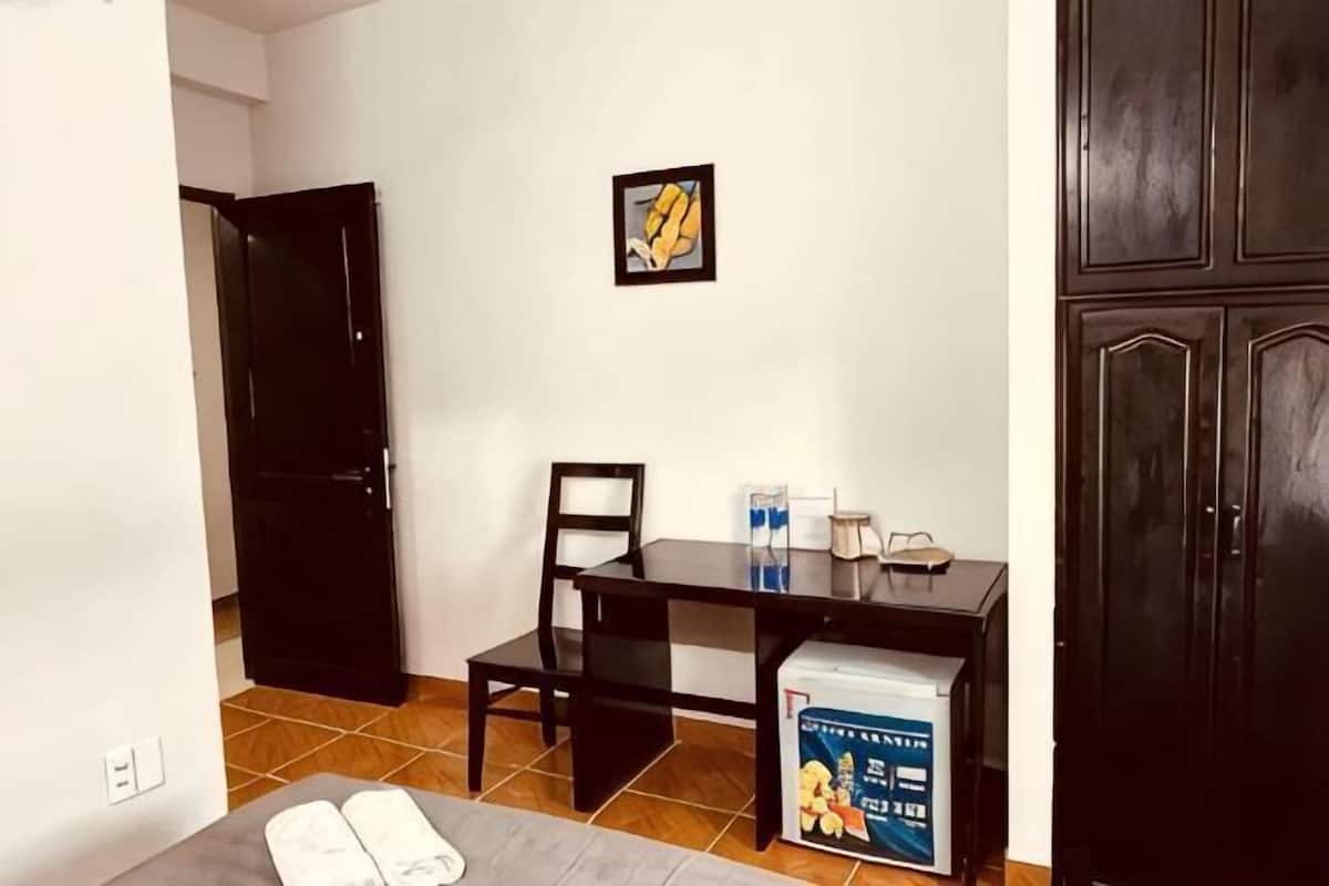 A small room with a bed, a table, a chair, a black cabinet, and a door.
