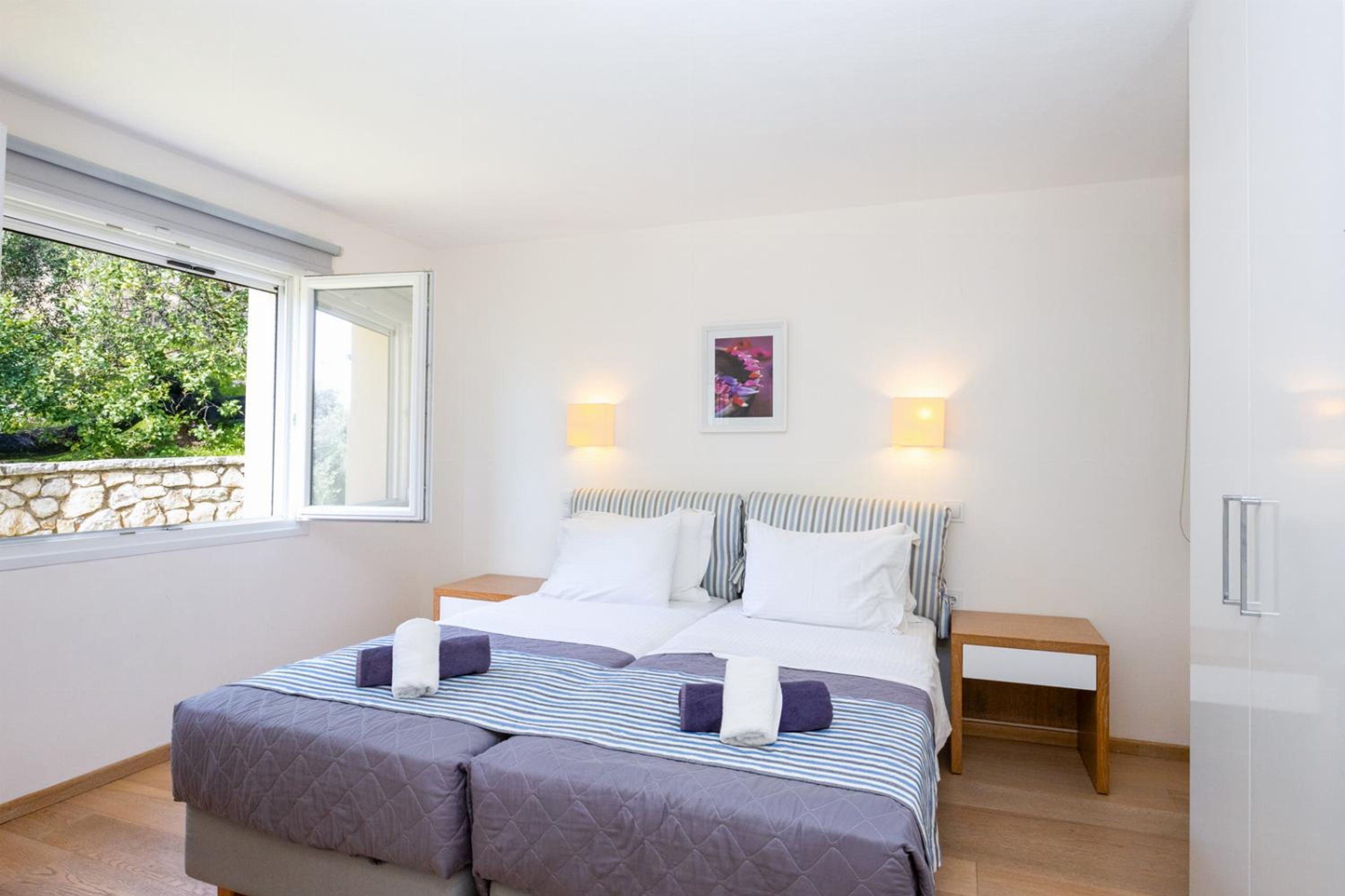 A double bed with striped bedding, two bedside tables, a wall-mounted light, and a window with a stone wall view.
