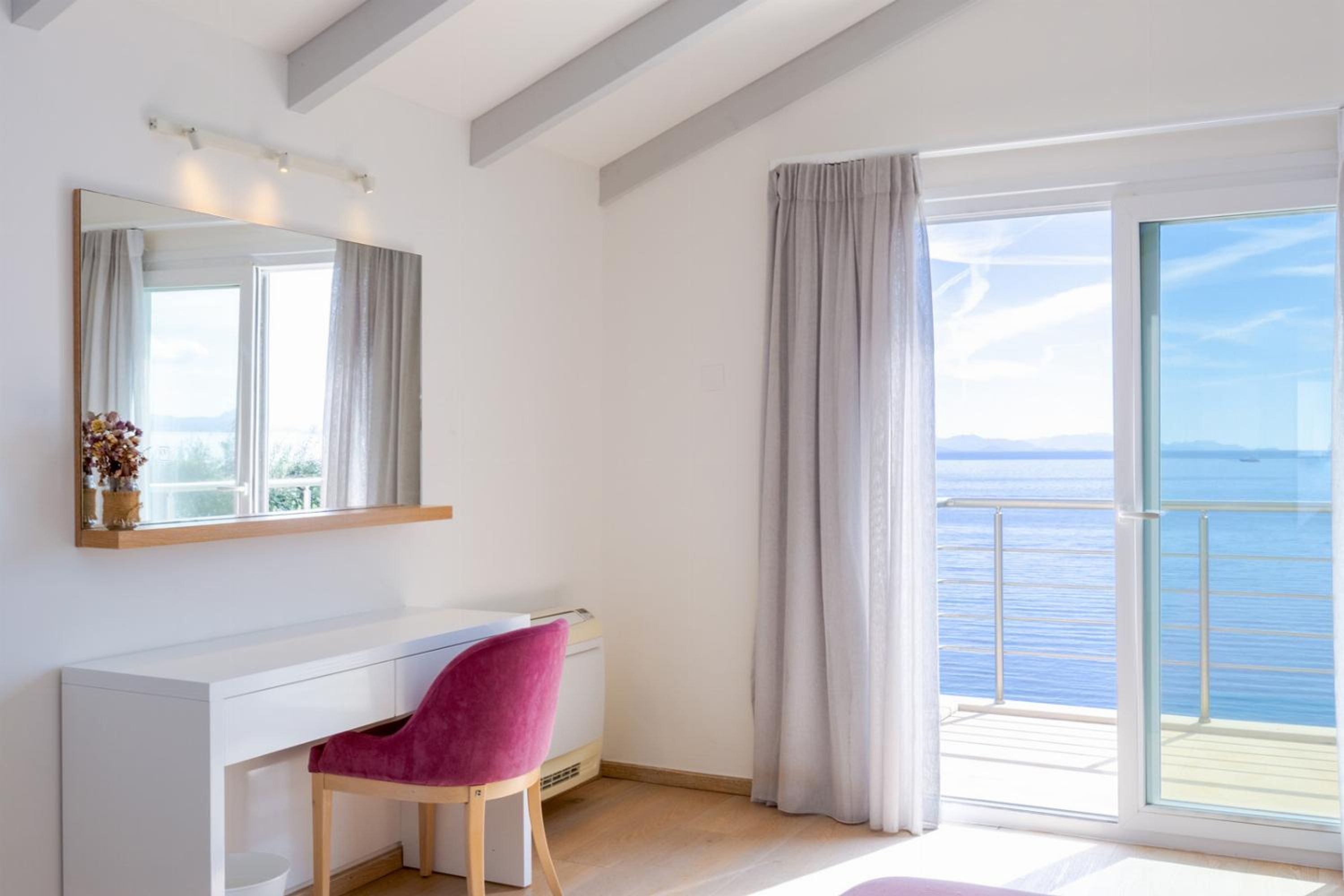 Una habitación moderna con un escritorio blanco, una silla rosa, un espejo grande y vistas al mar desde un balcón.
