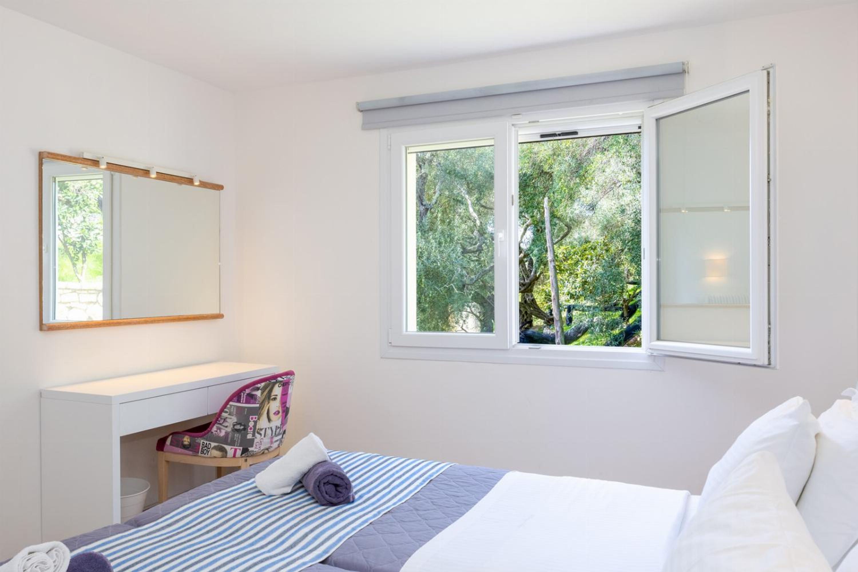 A bedroom with a bed, a desk, a chair, a window with a view of trees, and a mirror on the wall.