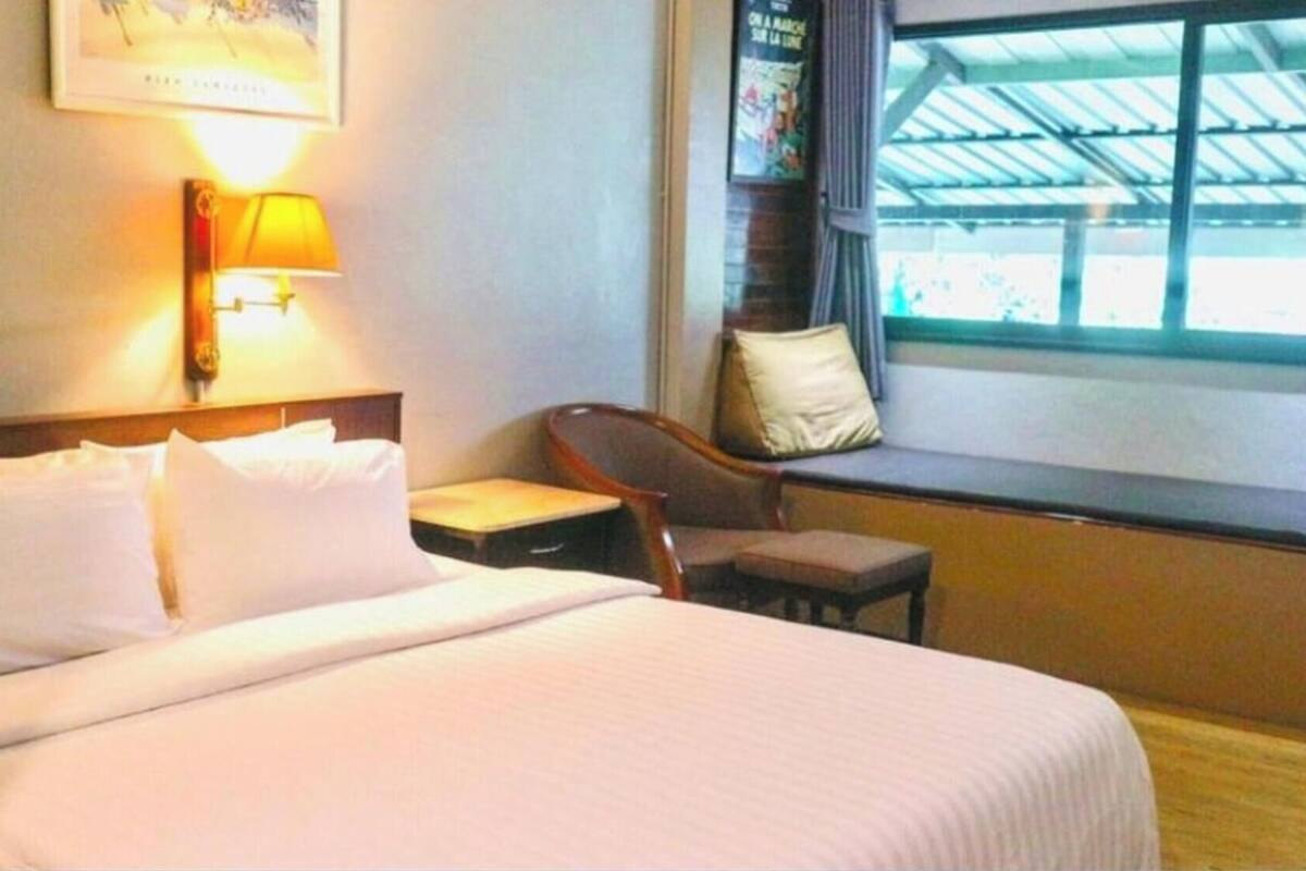A hotel room with a bed, a nightstand, a chair, a window, and a wall-mounted lamp.