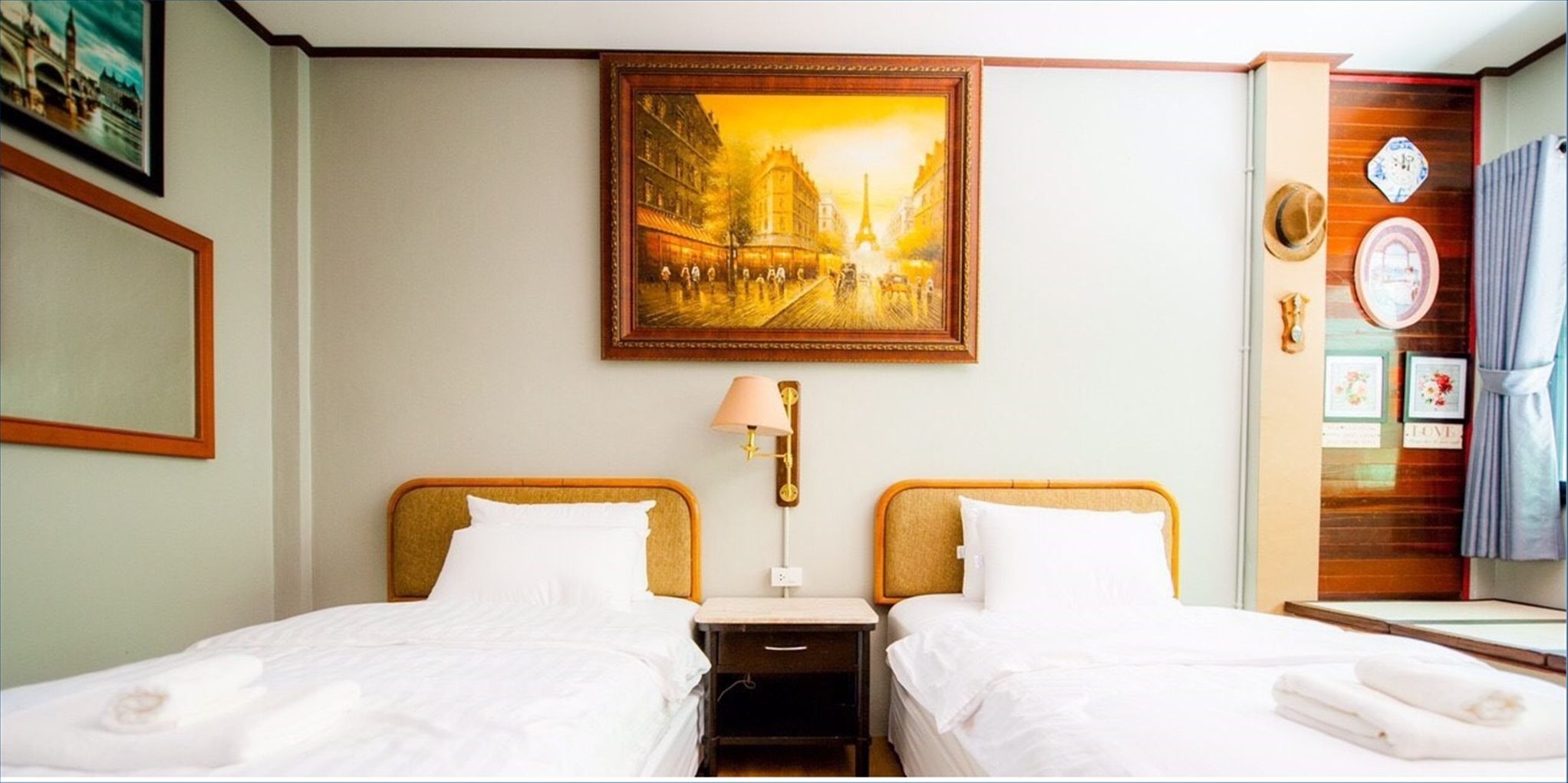 Une chambre d’hôtel avec deux lits, une table de chevet, une lampe, un tableau représentant un paysage urbain et un panneau en bois orné d’une horloge et d’objets décoratifs.
