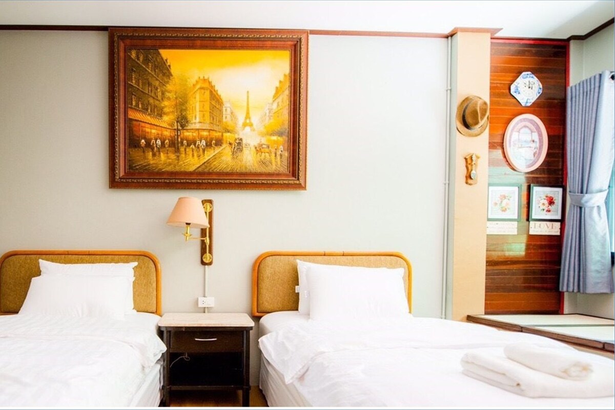 A hotel room with two beds, a nightstand, a lamp, a painting of a cityscape, and a wooden panel with a clock and decorative items.