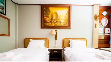 Une chambre d’hôtel avec deux lits, une table de chevet, une lampe, un tableau représentant un paysage urbain et un panneau en bois orné d’une horloge et d’objets décoratifs.