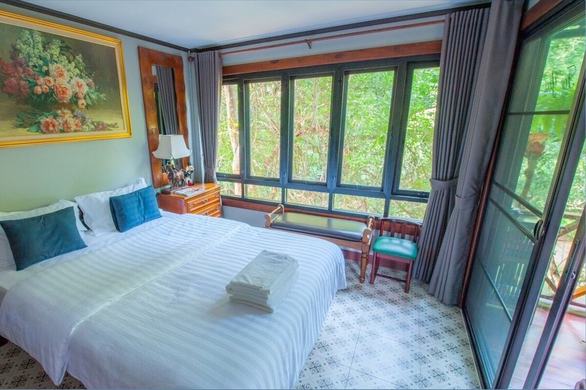 A hotel room with a large bed, a desk, a chair, a painting, and a window with a view of greenery.