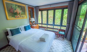 A hotel room with a large bed, a desk, a chair, a painting, and a window with a view of greenery. - Ban Mai Vintage (Pak Chong)