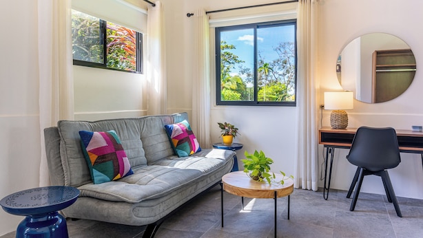 A modern living room with a grey sofa, colorful cushions, a small round wooden table, a black chair, a desk with a lamp, and a large window with curtains. - Casa Amor, 1-bedroom Villa with salt water pool, garden & mountain views (Ojochal)