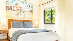 A bedroom with a wooden bed, two pillows, a nightstand with a lamp, a window with curtains, and a wall with a poster. - Casa Amor, 1-bedroom Villa with salt water pool, garden & mountain views (Ojochal)