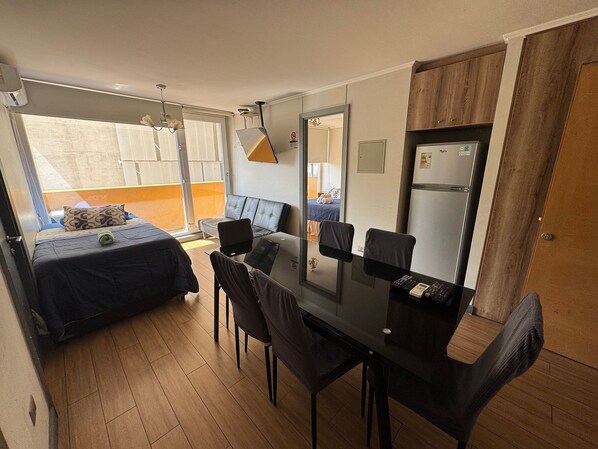 A compact room with a bed, a dining table with chairs, a refrigerator, and a window with a view. - EDIFICIO BARCELONA (Temuco)
