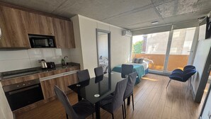 A modern kitchen with wooden cabinets, a microwave, and a sink. There is a dining area with a glass table and chairs. - EDIFICIO BARCELONA  (Temuco)