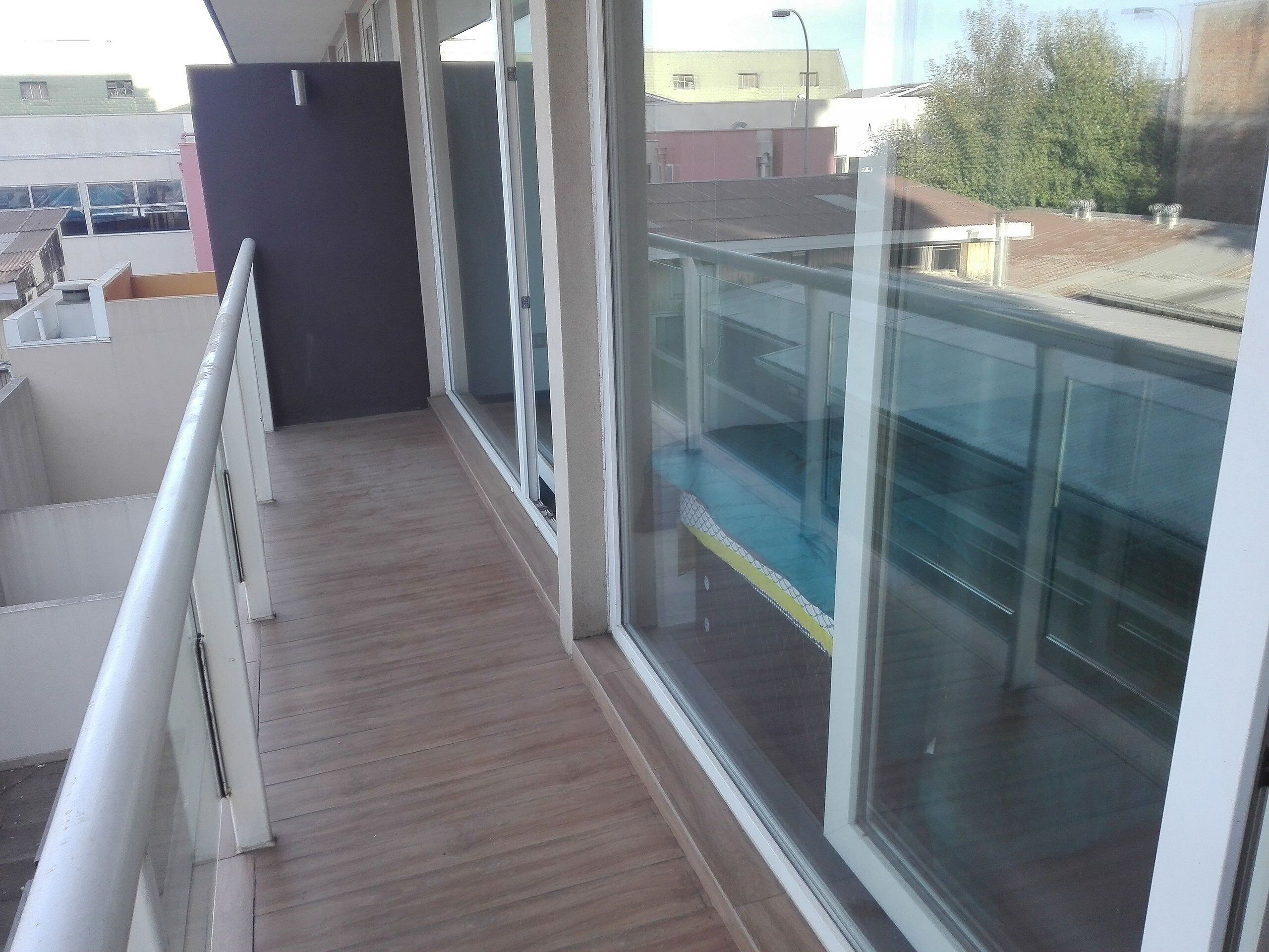 Un balcón con piso de madera, barandillas de vidrio y vistas a edificios y árboles.