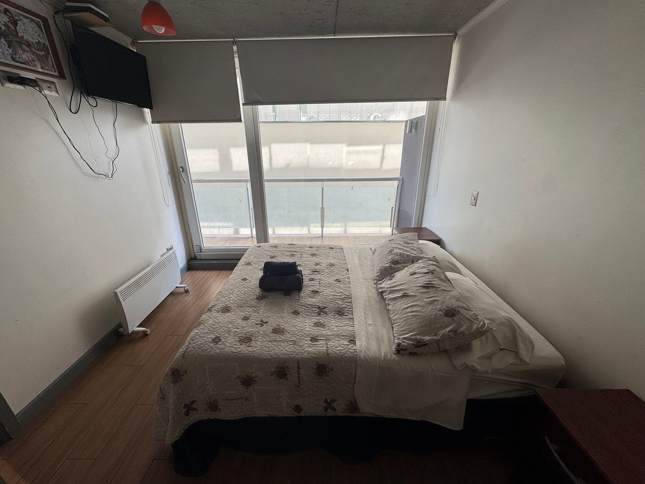 A bedroom with a bed, a radiator, a window with a view, and a TV mounted on the wall.
