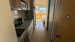 A narrow hallway with a kitchen area on one side, a door on the other, and a balcony with a view of buildings and trees. - EDIFICIO BARCELONA  (Temuco)