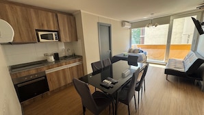 A modern kitchen with wooden cabinets, a built-in microwave, and a dining area with a glass table and chairs. - EDIFICIO BARCELONA  (Temuco)