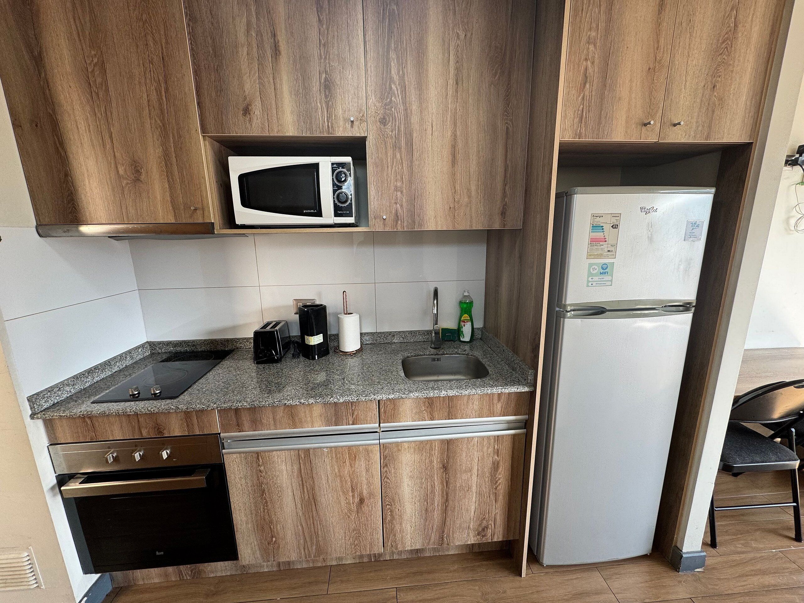 A compact kitchen with wooden cabinets, a microwave, a stainless steel refrigerator, a sink, and a stove.