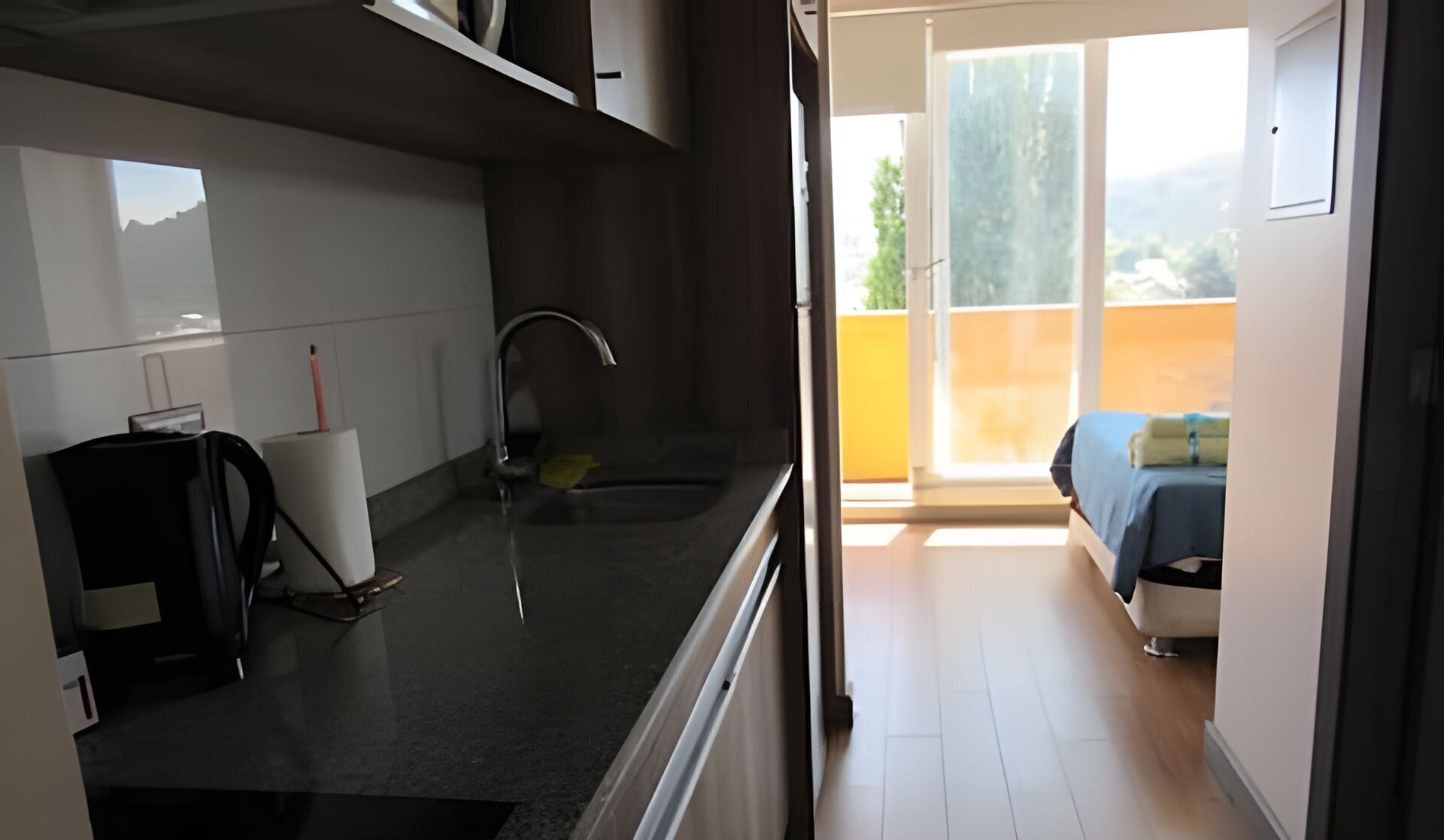 A compact kitchen with a sink, a small countertop, and a chair. A balcony with a bed and a view of the outdoors is visible through an open door.