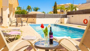A poolside area with lounge chairs, a table with a bottle of wine and flowers, and a clear blue sky. - Villa Solis With 3 Bedrooms and 3 Bathrooms (Paralimni)
