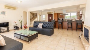 A living room with a sofa, coffee table, and TV. There is a kitchen area with a bar and stools. - Villa Solis With 3 Bedrooms and 3 Bathrooms (Paralimni)
