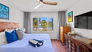 A hotel room with a bed, a ceiling fan, a window with a beach view, a TV, and a wooden dresser.