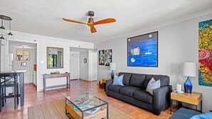 A modern living room with a glass coffee table, a dark sofa, a wooden side table with a lamp, and a ceiling fan.