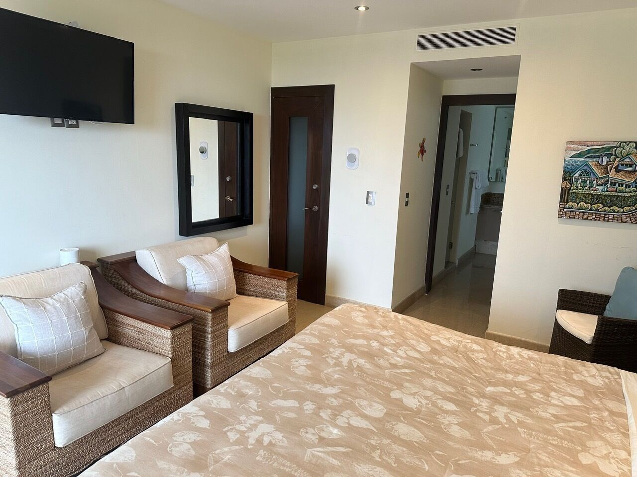 A hotel room with a large bed, two wicker sofas, a flat-screen TV, and a door leading to another room.