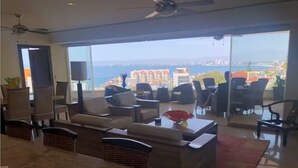 Un'ampia zona giorno con un divano grande, un tavolino da caffè e un tavolo da pranzo. Si gode di vista sull'oceano e sugli edifici sullo sfondo.