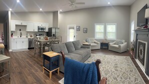 Ein geräumiges Wohnzimmer mit grauem Sofa, zwei Sesseln, einem Couchtisch und einem blauen Sessel. Es gibt eine Küchenzeile mit weißen Schränken und einen Esstisch. Das Zimmer verfügt über große Fenster und einen Deckenventilator.