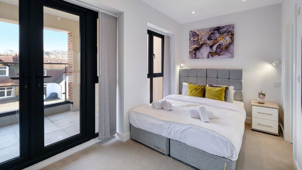 A modern bedroom with a large bed, a grey headboard, a small bedside table with a lamp, and a view of the outside through a sliding door.