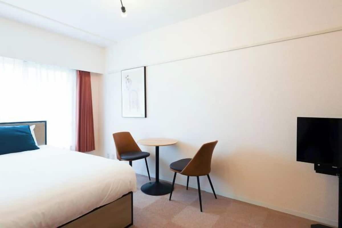 Une chambre d’hôtel avec un lit, une petite table ronde et deux chaises, un téléviseur et une fenêtre avec des rideaux.