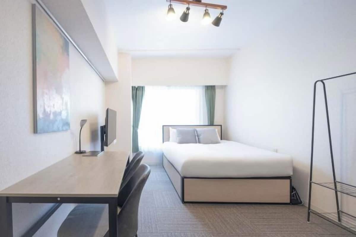 Une chambre d’hôtel avec un lit, un bureau, une chaise, une télévision et une fenêtre avec des rideaux.