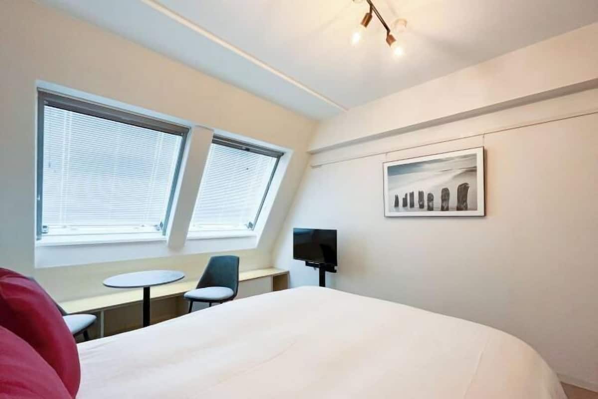 Ein Hotelzimmer mit einem Bett, zwei Fenstern, einem Schreibtisch mit Stuhl, einem Fernseher und einem gerahmten Bild an der Wand.