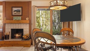 A cozy living room with a fireplace, a round wooden dining table, and chairs. A television is mounted on the wall. - Outpost: Larkspur 1212 (Wilson)