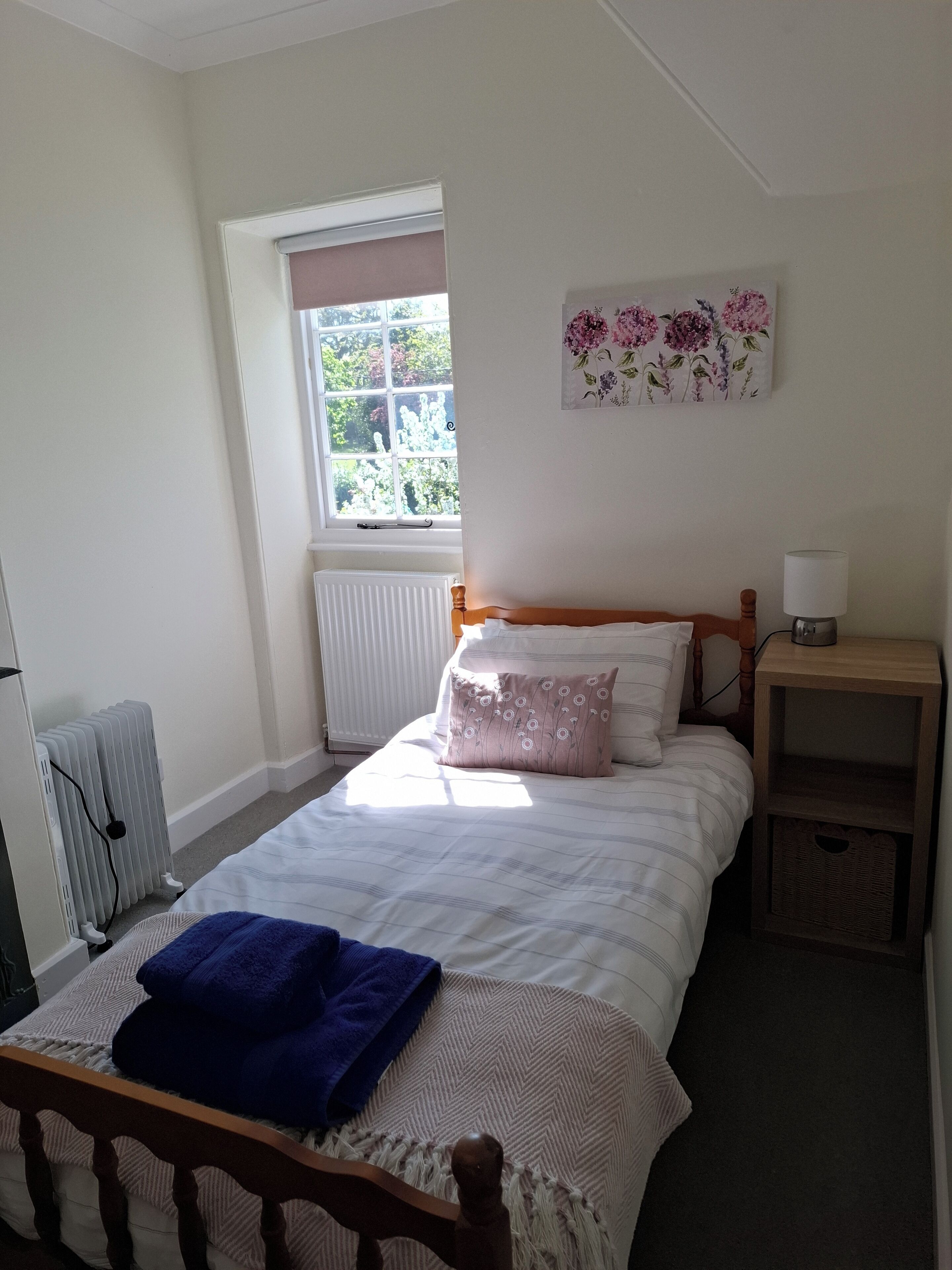 A bedroom with a bed, a radiator, a window, a wall-mounted lamp, and a small bedside table.