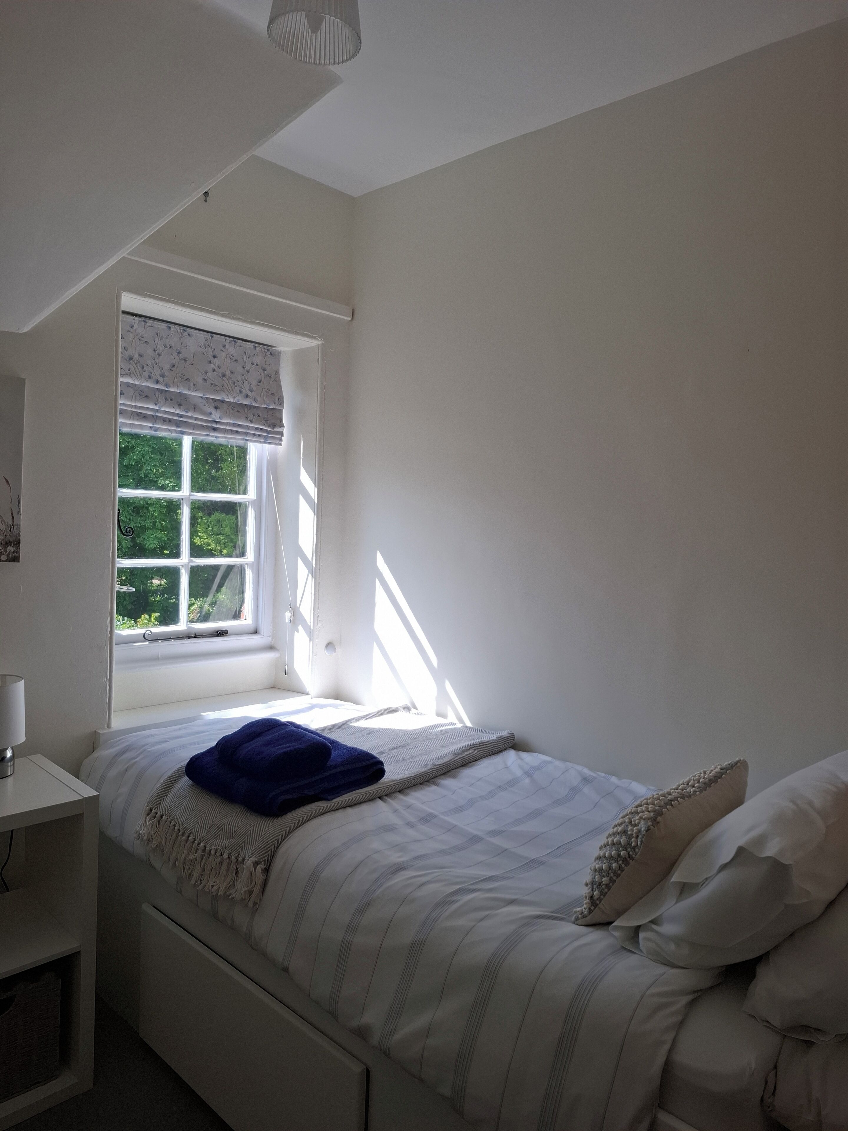 A bedroom with a bed, a window with a view of greenery, and a bedside table with a lamp.