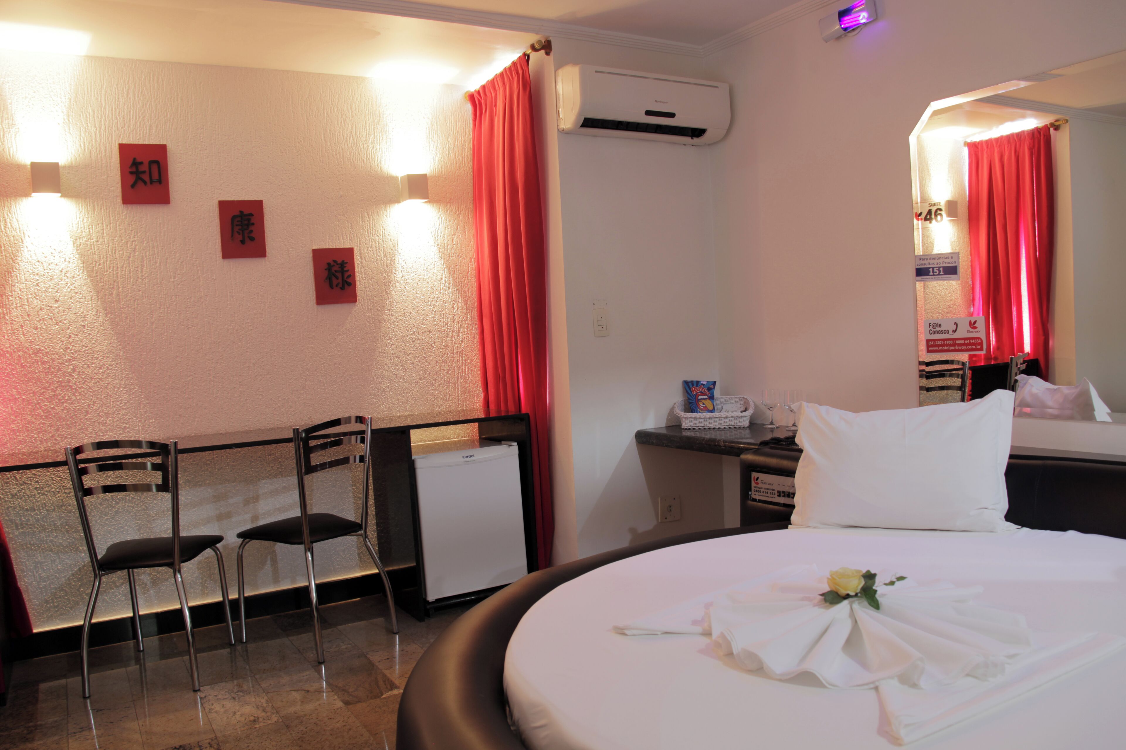 Quarto de hotel com cama, duas cadeiras, uma mesa pequena, um refrigerador e um espelho.