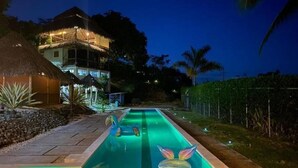 Une piscine dotée d’une structure surmontée d’un toit de chaume, avec des palmiers en arrière-plan.