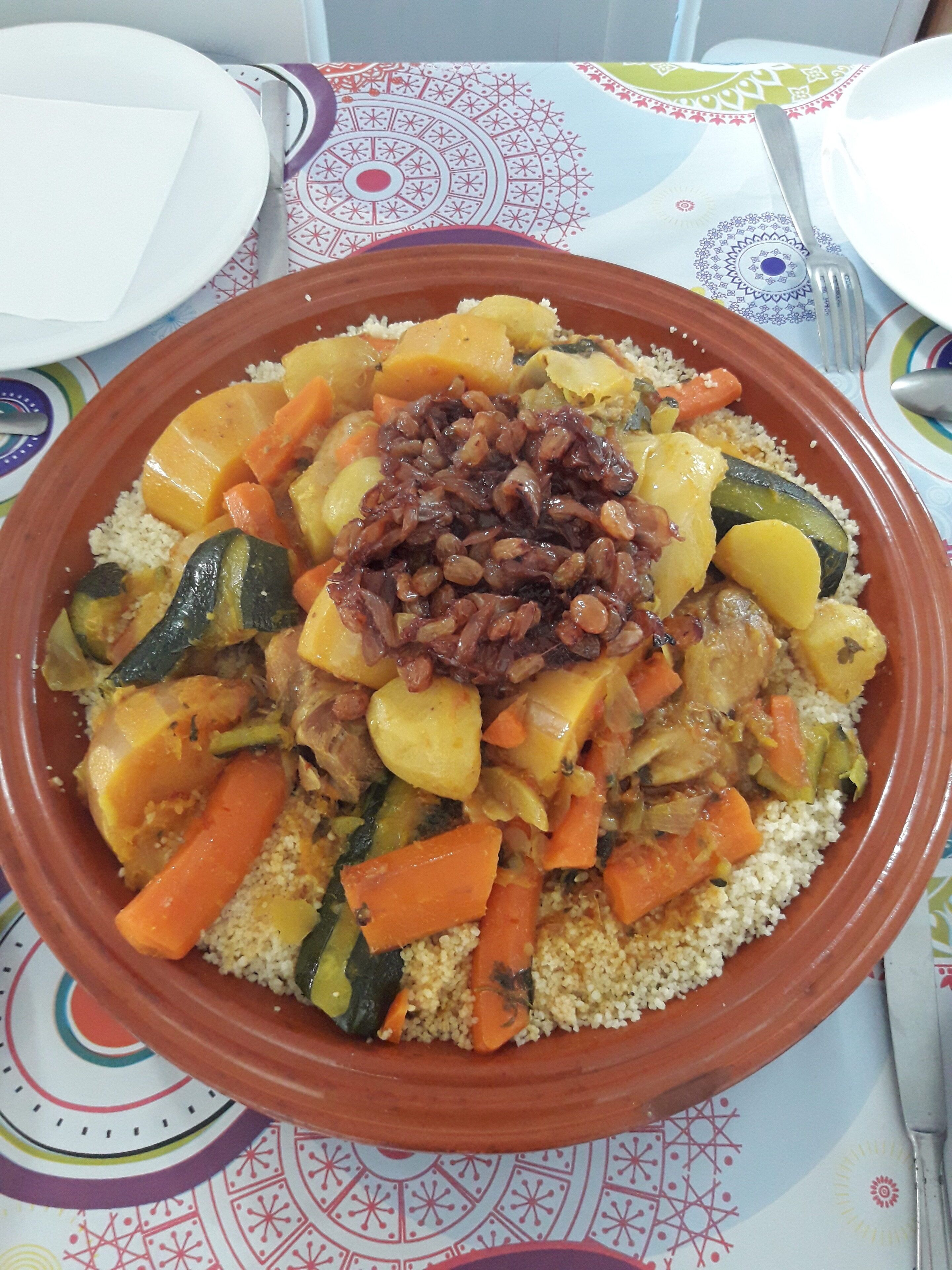 Un plato grande y redondo de terracota lleno de cuscús, verduras y un adorno de frutas secas, sobre un mantel estampado.