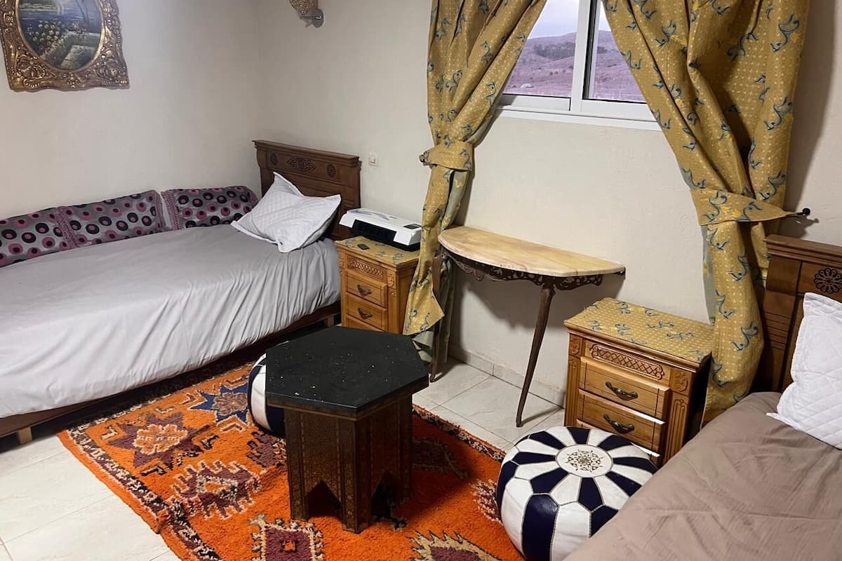 A bedroom with a bed, a small table, a nightstand, a chair, and a window with curtains.