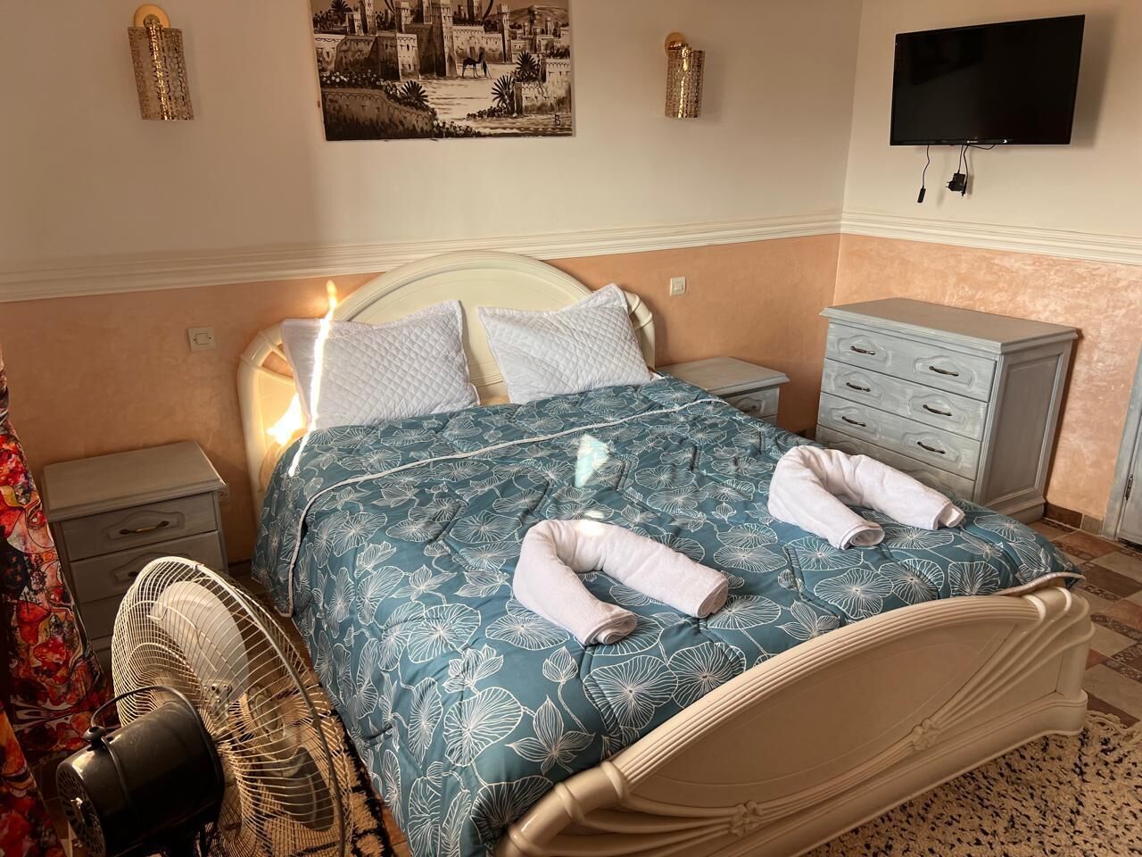 Una cama bien hecha con cubrecama estampado con motivos florales, dos almohadas y dos toallas. Se aprecia una mesita de noche con una lámpara, un cajón pequeño y un televisor montado en la pared.