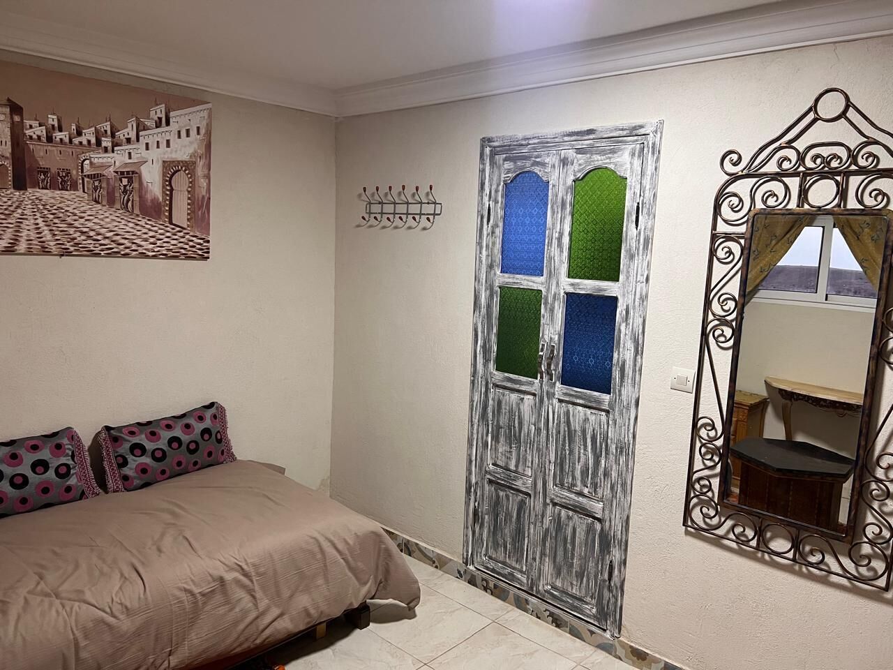 A bedroom with a bed, a door with a green window, a wall-mounted mirror, and a wall art piece.