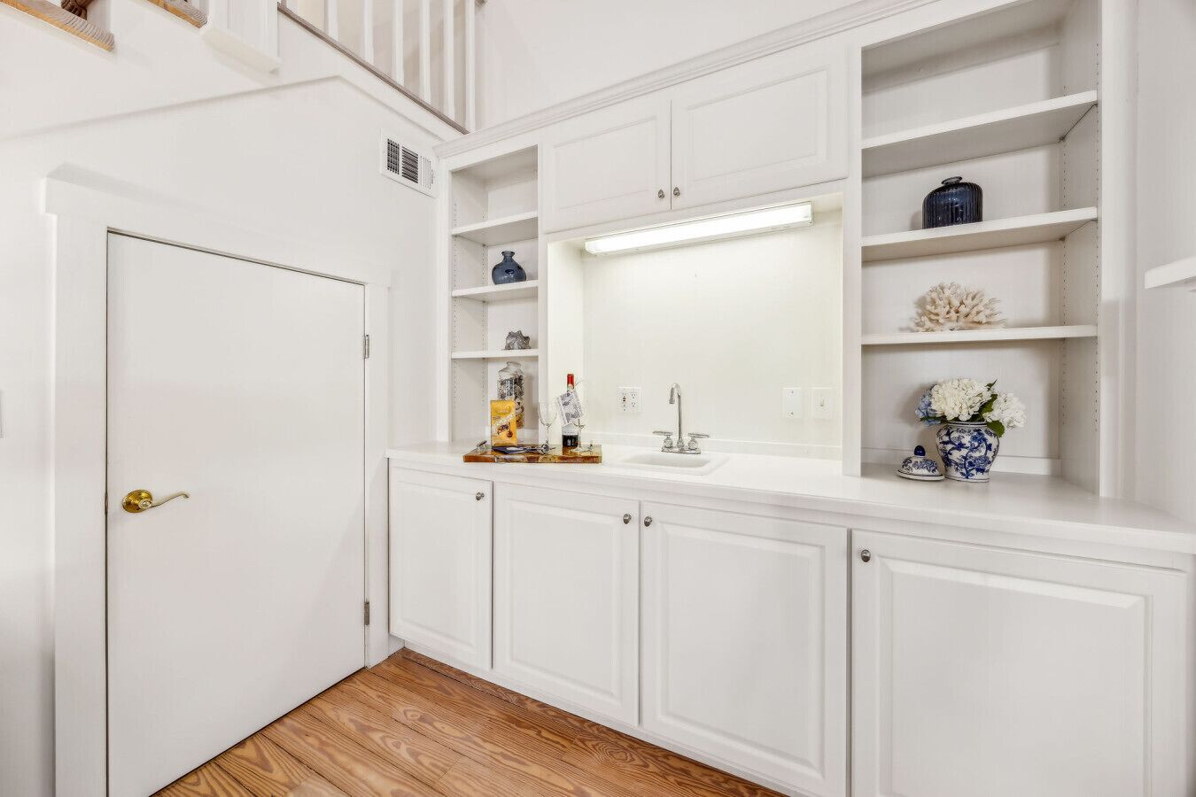 A white bathroom with a wooden floor, white cabinets, a mirror, and a shelf with decorative items.