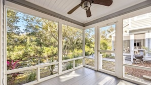 Un espacio exterior techado con ventilador de techo, ventanas amplias y vistas a un jardín y una casa.