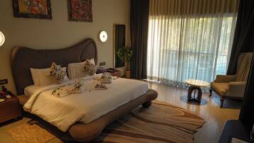 Una cama grande con cabecera, una mesita de noche, una pequeña mesa redonda, una silla y un amplio ventanal con cortinas.