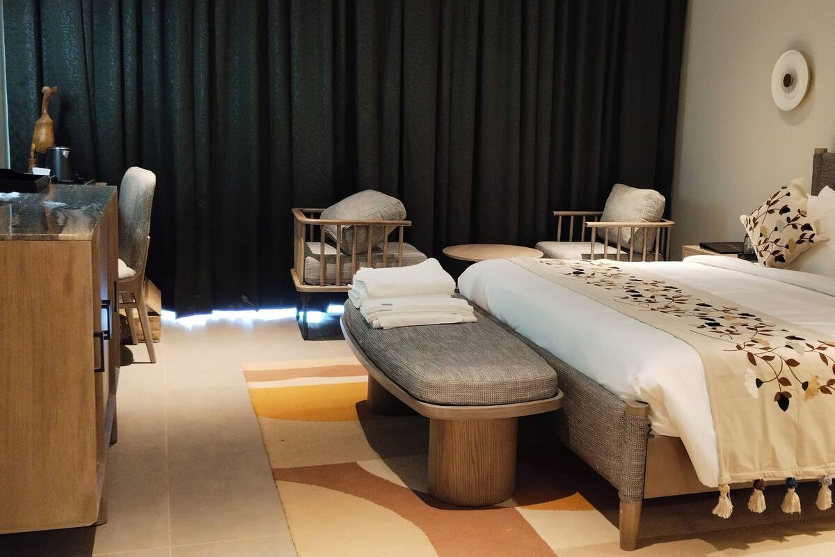 A modern hotel room with a large bed, a wooden desk, a bench, and a small table.