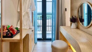 A hotel room with a wooden vanity, a round mirror, a small stool, a wardrobe with hanging clothes, and a view through a glass door. - Maison Villa By The Sea (Ho Chi Minh City)