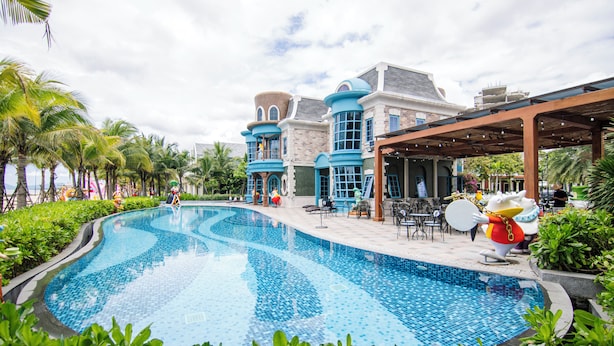 A large swimming pool with a curved design, surrounded by lush greenery and palm trees. - Maison Villa By The Sea (Ho Chi Minh City)